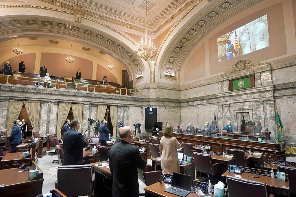 Senate members stand as a ceremonial presentation of colors is done virtually on a video screen above on Monday at the Capitol in Olympia. Washington states Legislature convened Monday under a large security presence because of concerns about efforts by armed groups who might try to disrupt the proceedings or occupy the Capitol, which is closed to the public due to the ongoing pandemic. (AP Photo/Ted S. Warren)